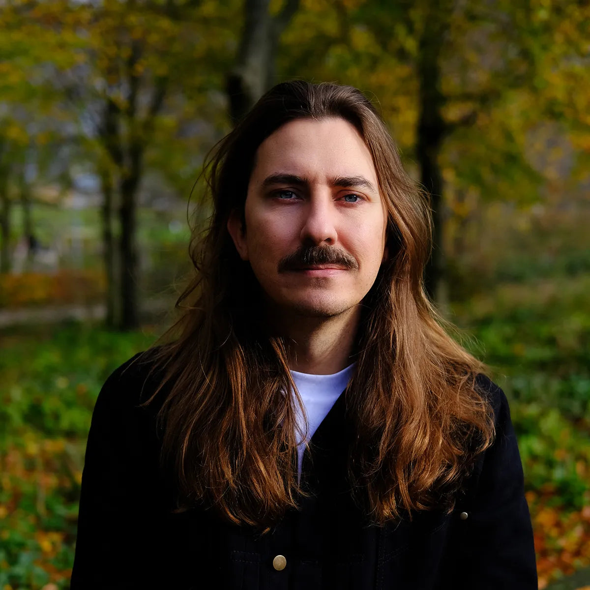 Man with long hair and a mustache standing in a forest during autumn.